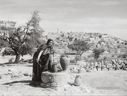 Bethlehem from south with woman figure, Sofie at the well, clad in richly embroidered costume and drawing water. Bethlehem, Palestine, 1938. Photographer unknown. Image courtesy http://www.photographium.com/